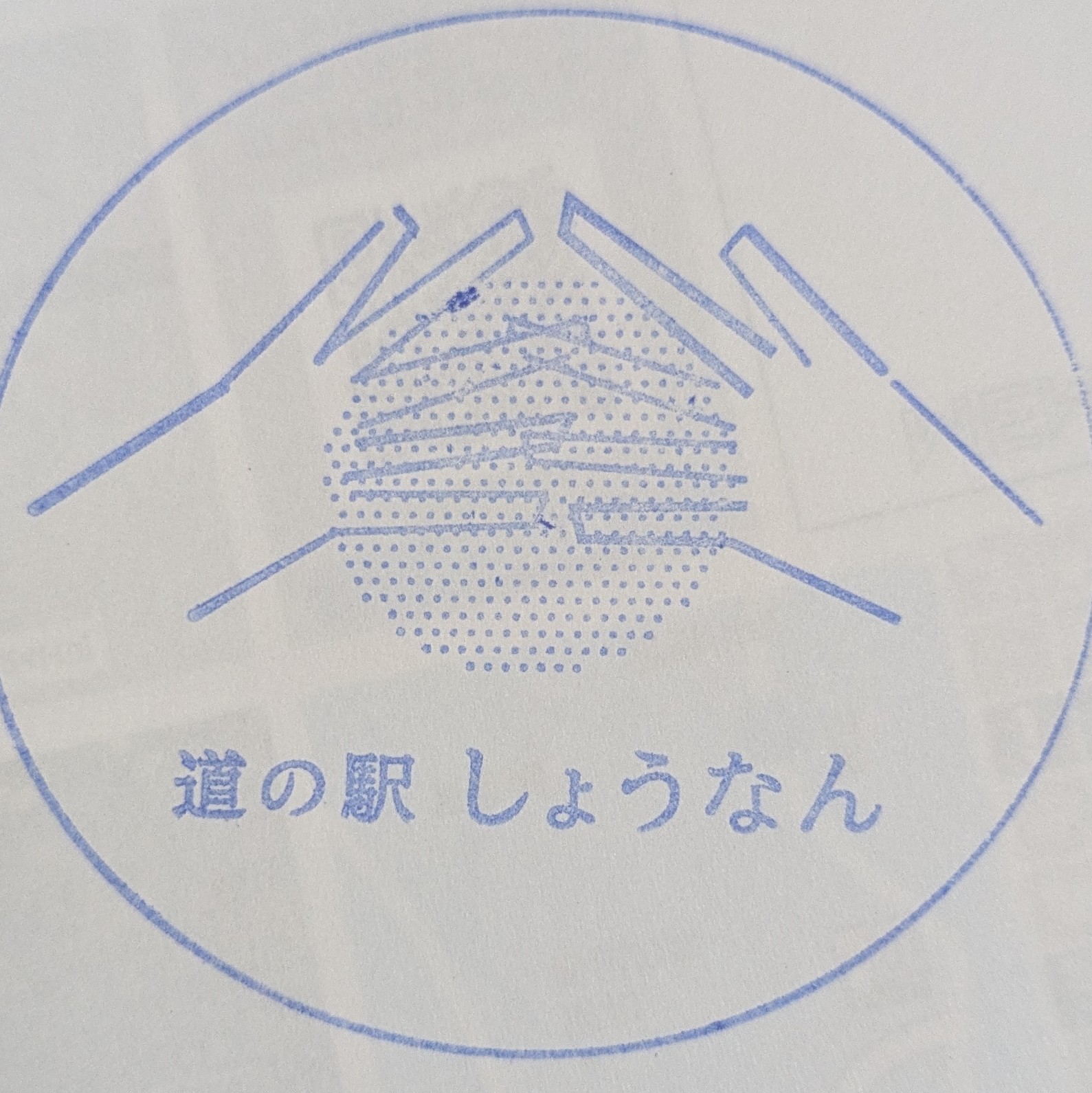 青いインクで押された円形のスタンプ。左右の手が指先を合わせて山形をつくり、その下の点描の円の中に三角屋根の建物が描かれている。下に「道の駅 しょうなん」の文字