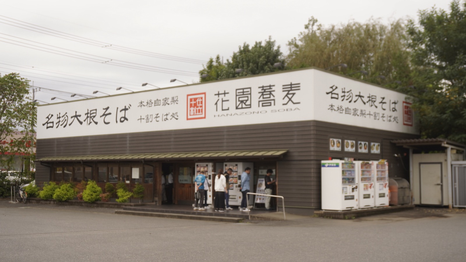 「花園蕎麦」「名物大根そば」と書かれた看板を掲げた平屋の店舗と、入口前の券売機に並ぶ人