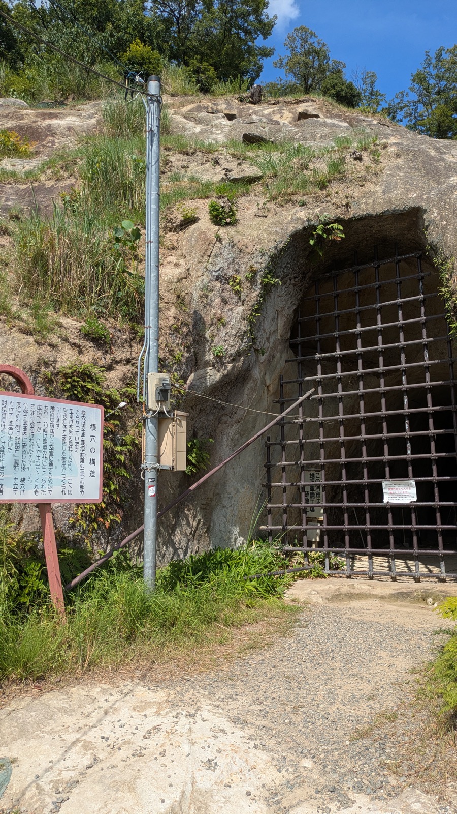岩肌に掘られたアーチ形のトンネルの入口が鉄の格子でふさがれ、左手に「横穴の構造」の説明板が立っている
