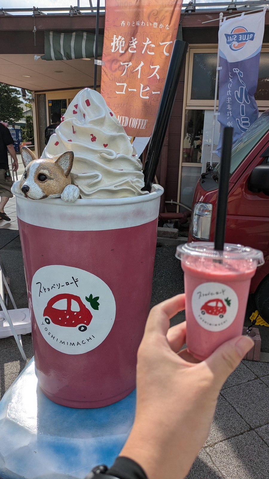 「ストロベリーロード YOSHIMIMACHI」のロゴが入ったピンクのドリンクを手に持ち、同じデザインの大きなカップ型オブジェと並べている