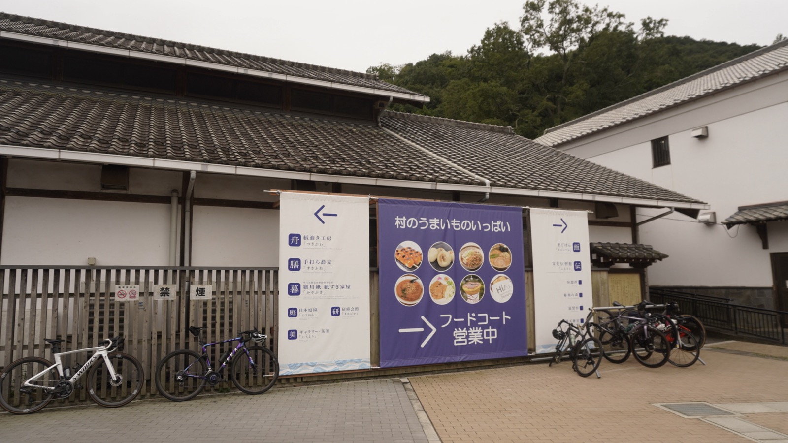 瓦屋根の建物の前に「村のうまいものいっぱい フードコート営業中」と書かれた紫の幕がかかり、その両脇の柵にロードバイクが何台も立てかけられている
