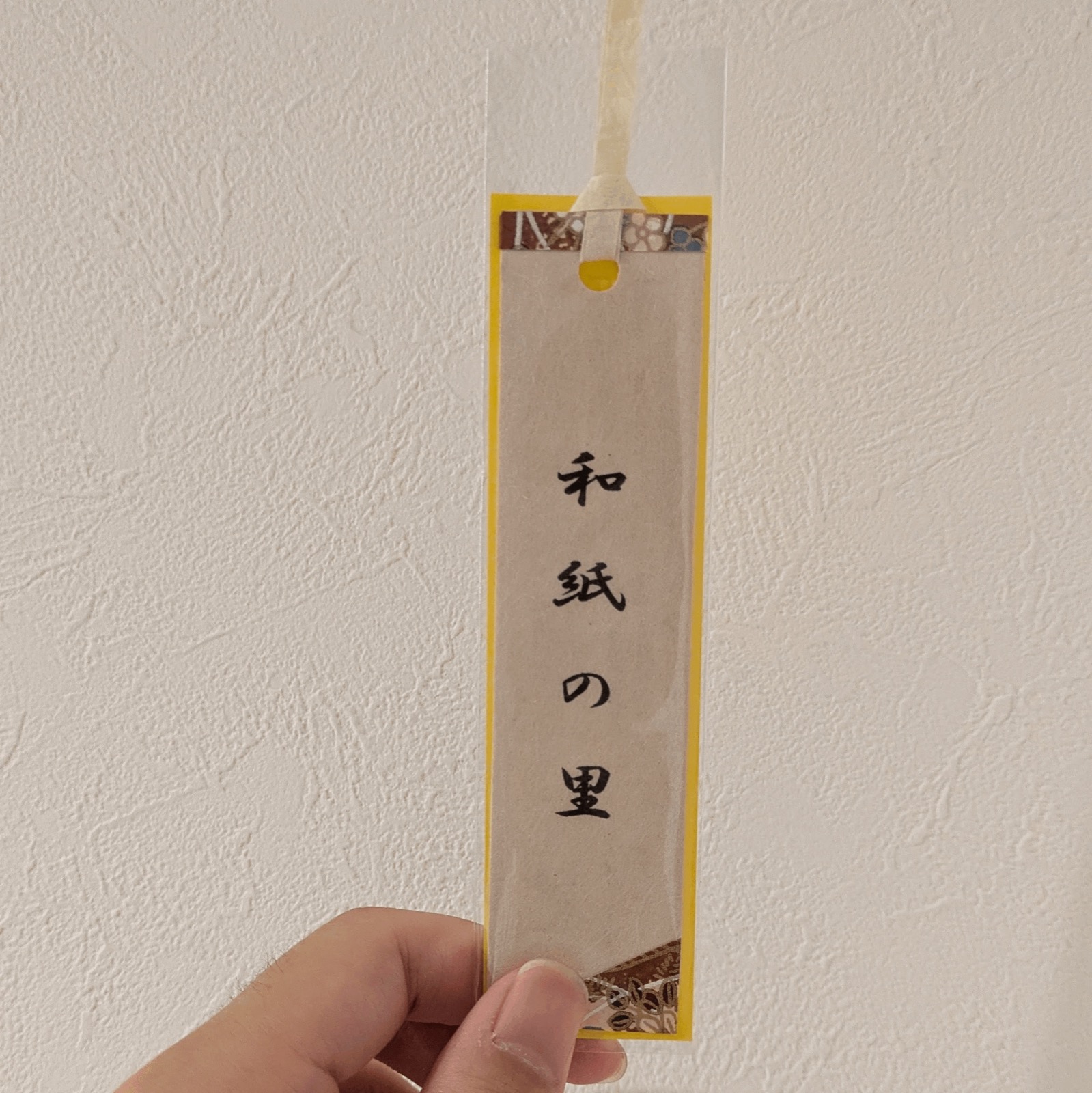 白い壁の前で手に持った縦長の栞。薄い和紙に筆文字で「和紙の里」と書かれ、黄色い縁と千代紙の柄、ひもが付いている
