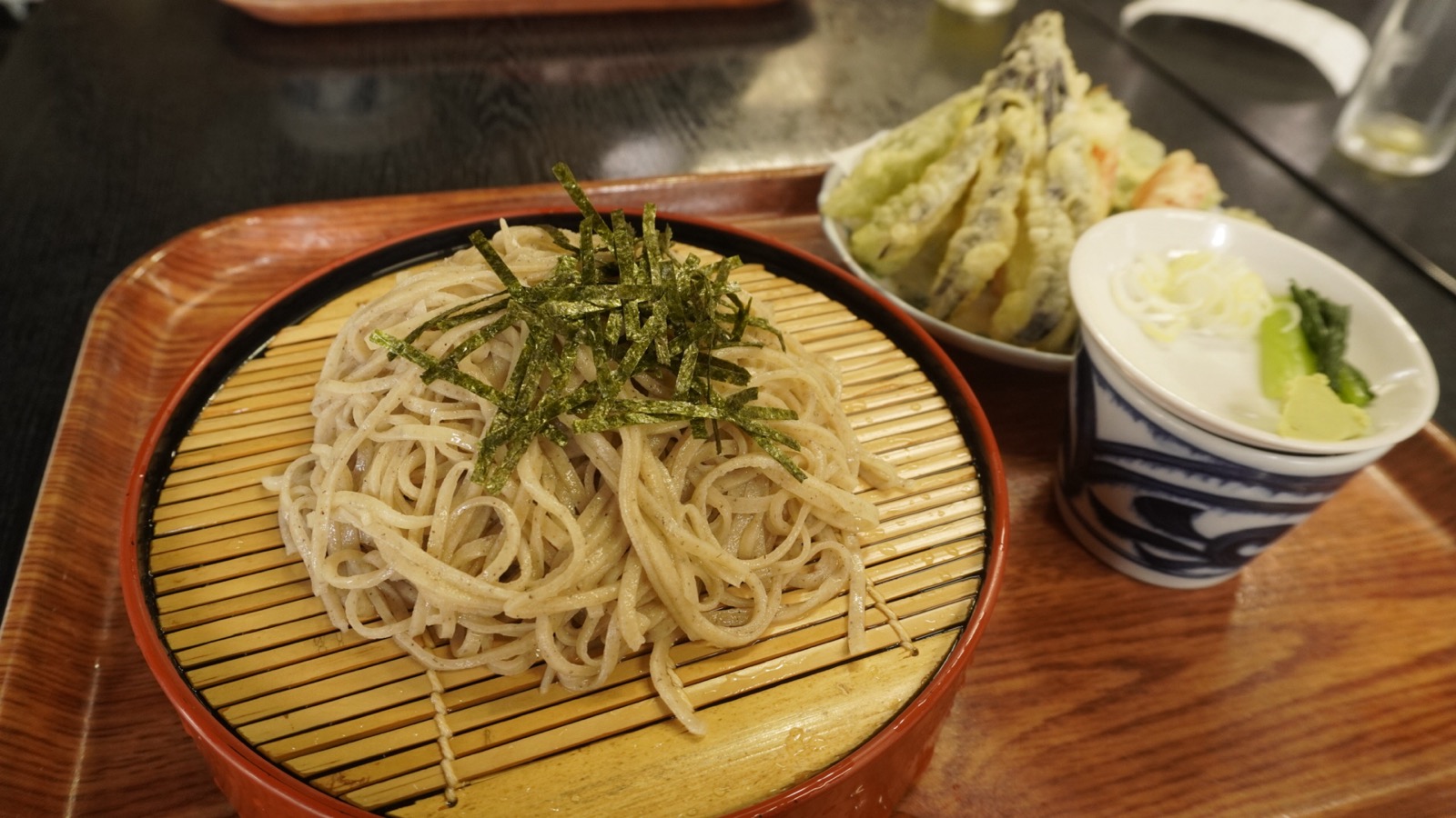 赤い縁の丸いせいろに盛られ、刻み海苔がのったざるそば。奥に野菜の天ぷらの皿、右に薬味の入った染付のそば猪口がある