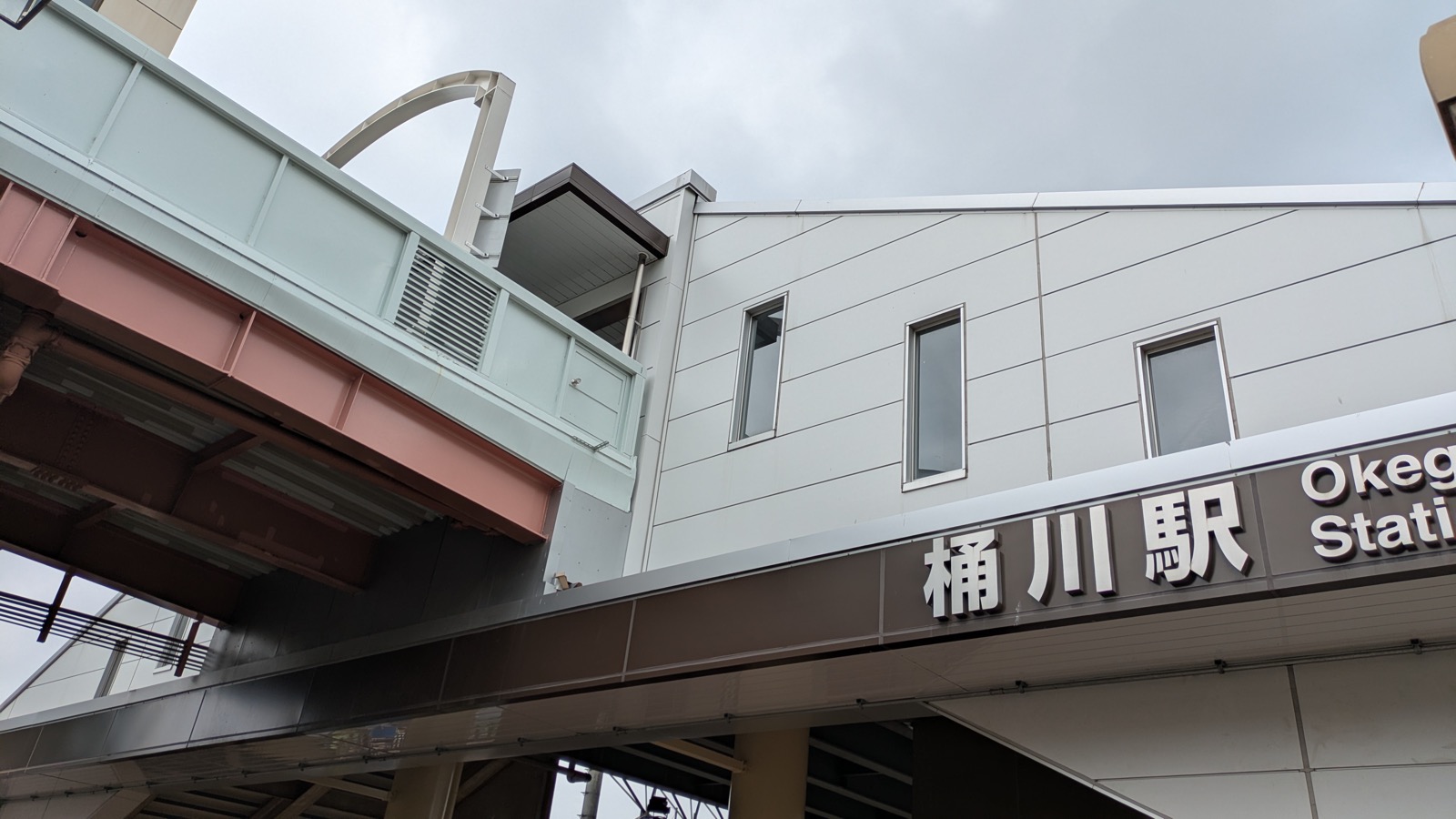 見上げるアングルで撮影した「桶川駅」の駅名標とホーム上家