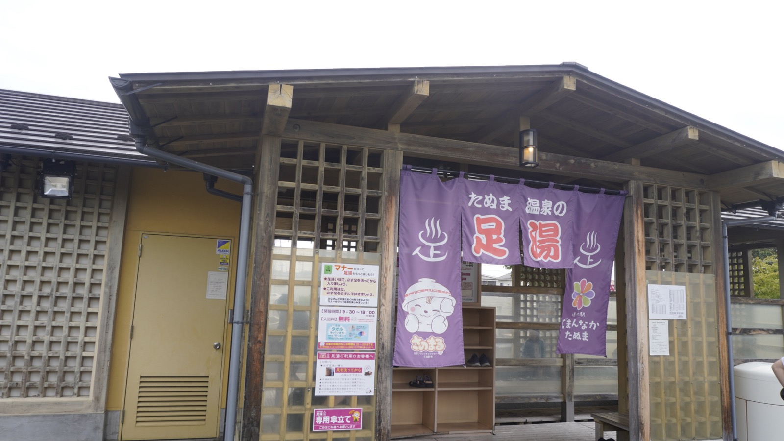 木の格子に囲まれた足湯の建物。「たぬま温泉の足湯」と書かれた紫ののれんがかかっている