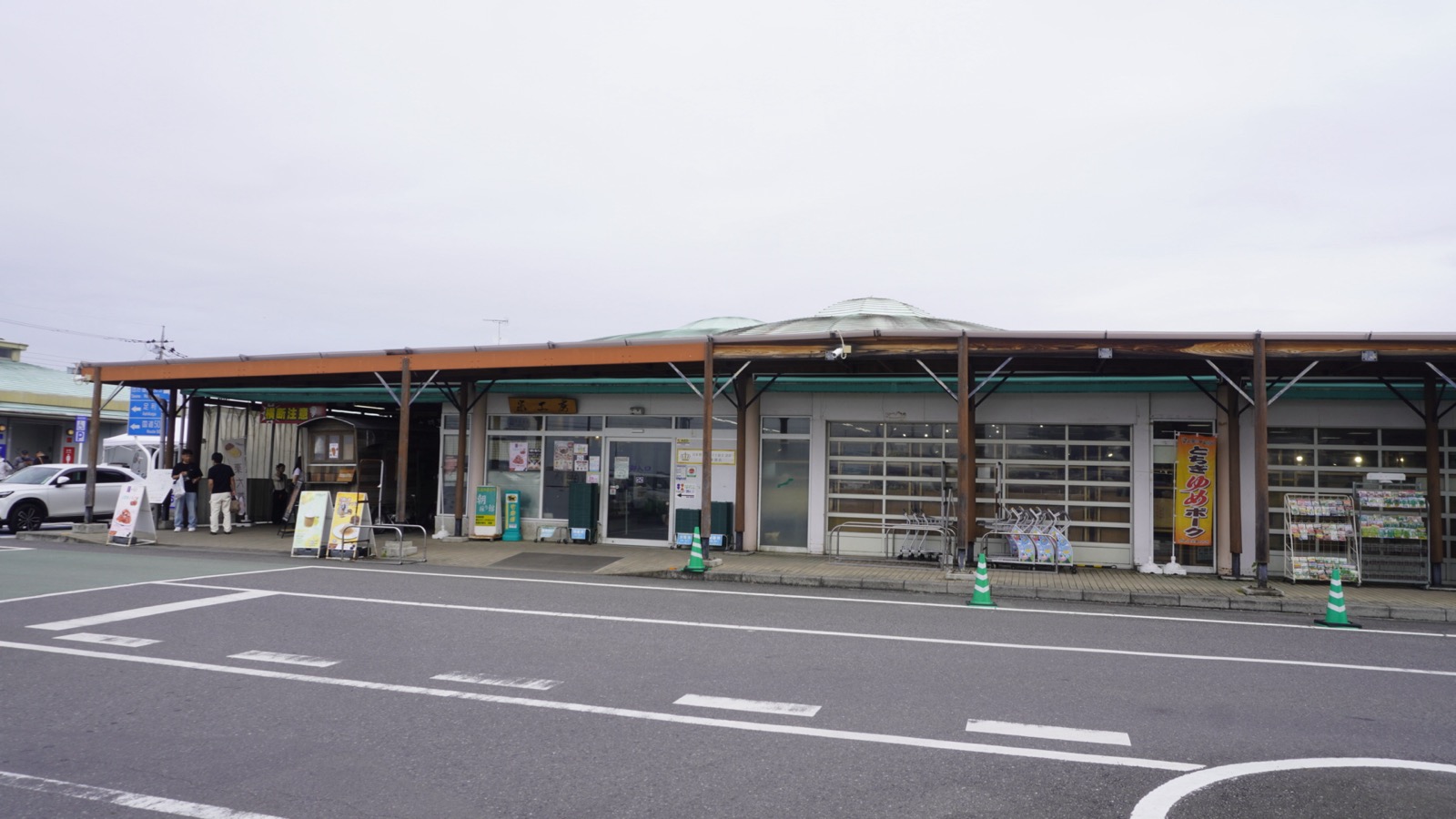 曇り空の下、駐車場から見た道の駅どまんなかたぬまの建物。木の柱と緑の屋根が横に長く続く