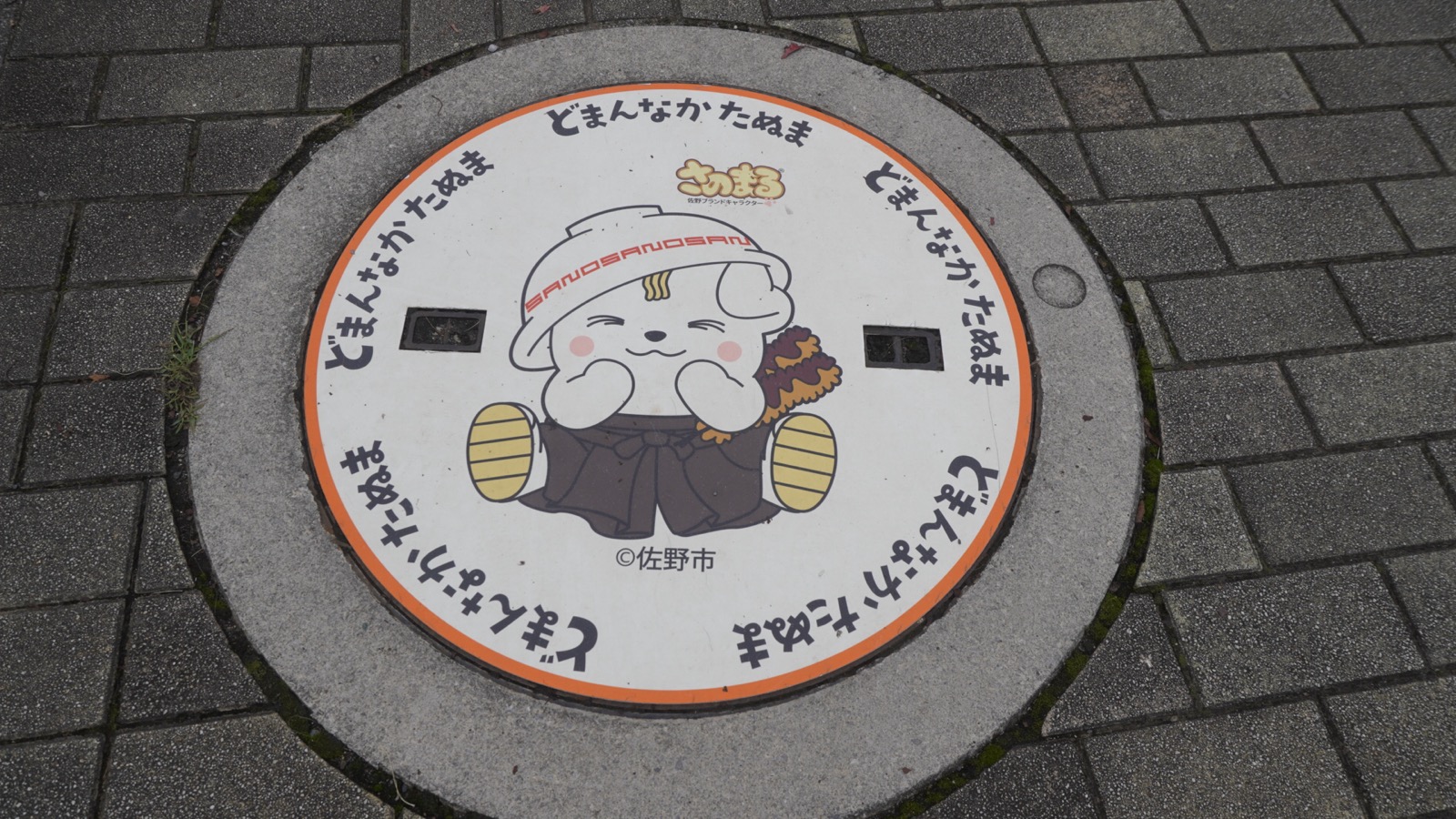 石畳の上にある、さのまるが描かれた白いマンホールの蓋