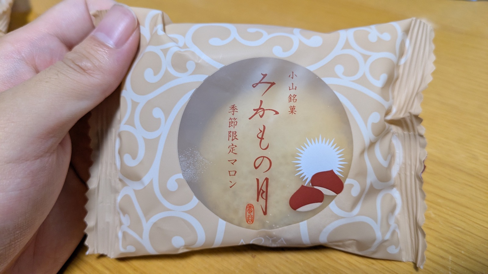 手に持った「みかもの月 季節限定マロン」の個包装。薄茶色の唐草模様の袋の丸窓から、黄色いカステラ生地がのぞき、栗のイラストが添えられている