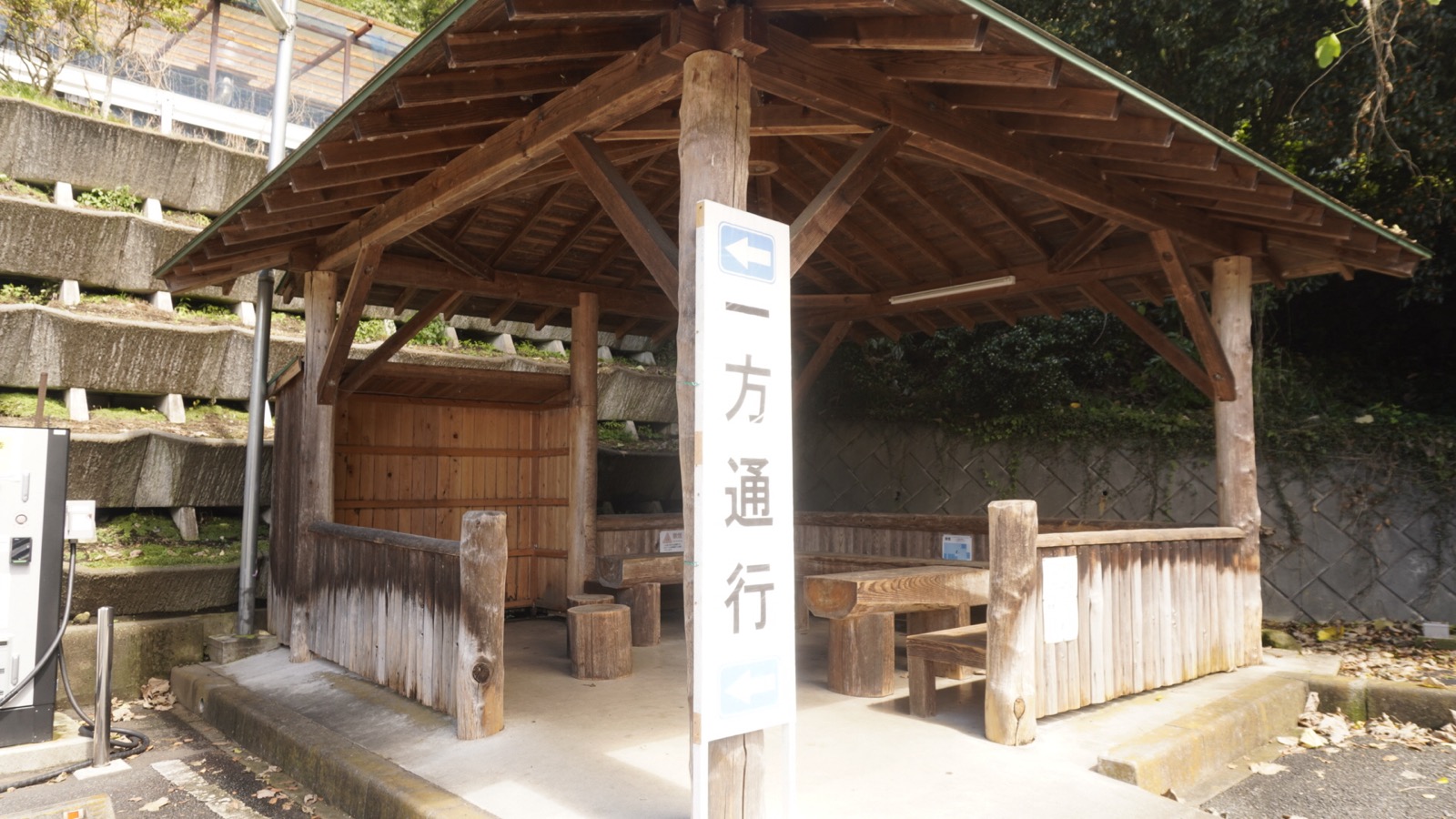 駐車場の一角にある、丸太の柱と木の屋根のあずまや。木のベンチとテーブルが見え、手前に「一方通行」の白い標識が立つ