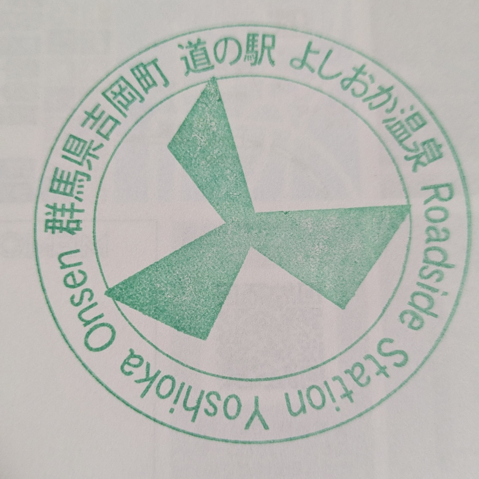 緑のインクで押された円形のスタンプ。中央に3枚羽根の風車、周りに「群馬県吉岡町 道の駅 よしおか温泉 Roadside Station Yoshioka Onsen」の文字がある