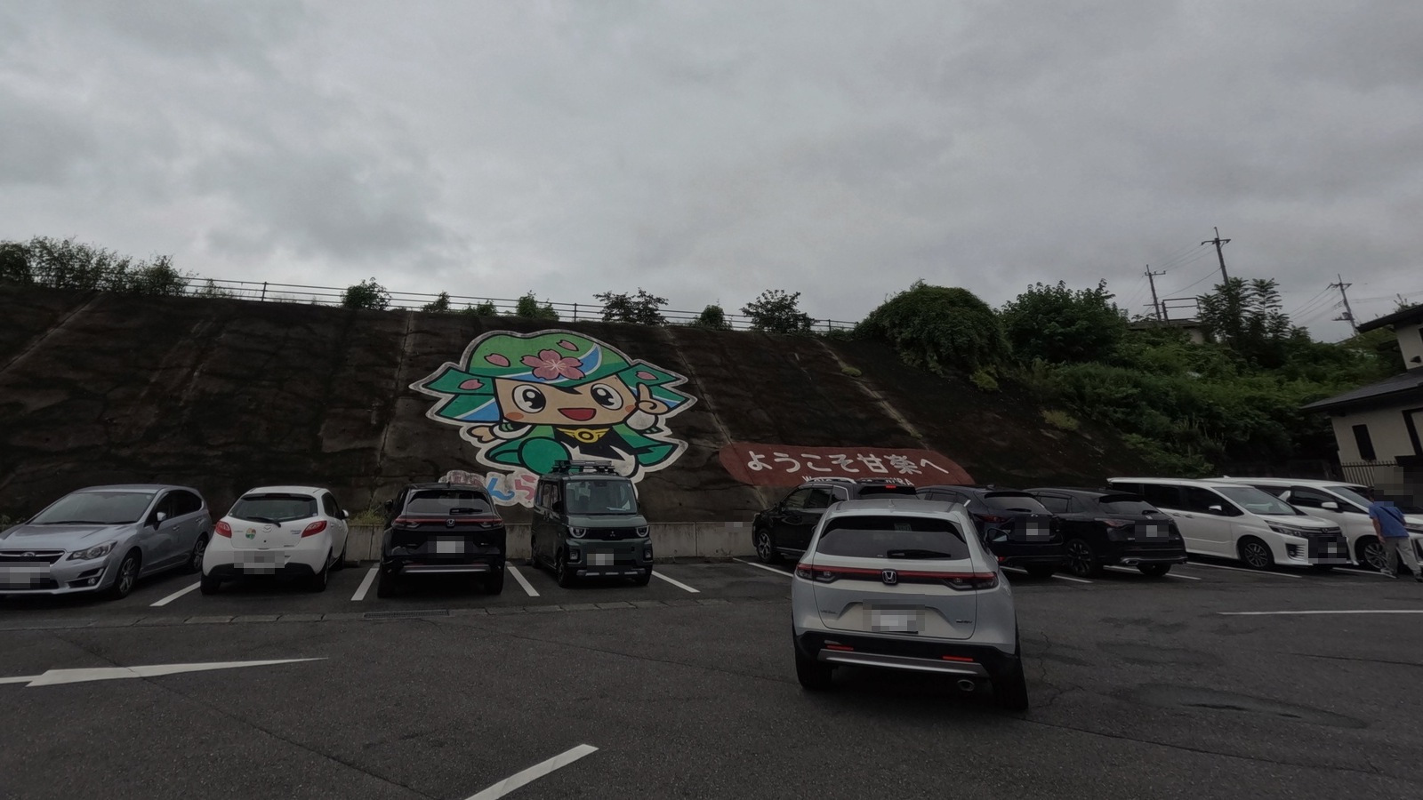 曇り空の下、駐車場の擁壁に描かれた、桜の花をあしらった緑色の鎧兜をかぶるキャラクターと、「ようこそ甘楽へ」と書かれた看板