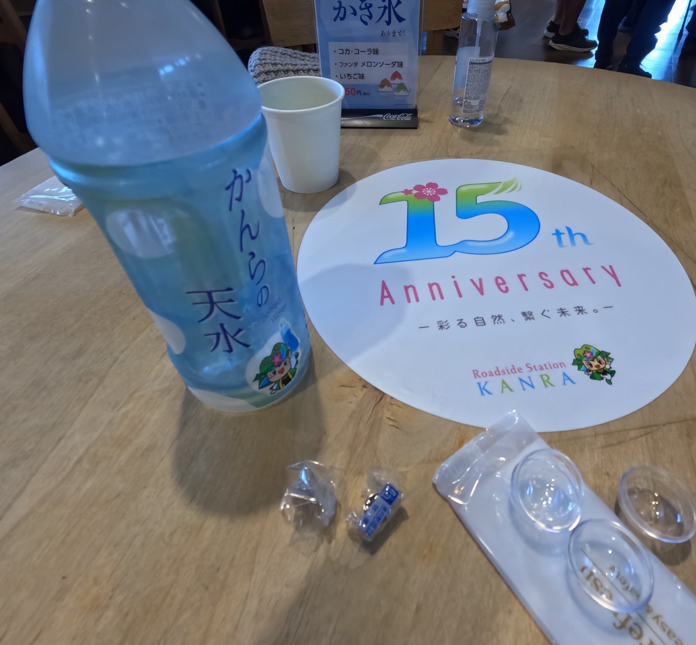 木のテーブルに置かれた「かんらの天水」のペットボトルと、15周年記念の白い楕円形の案内