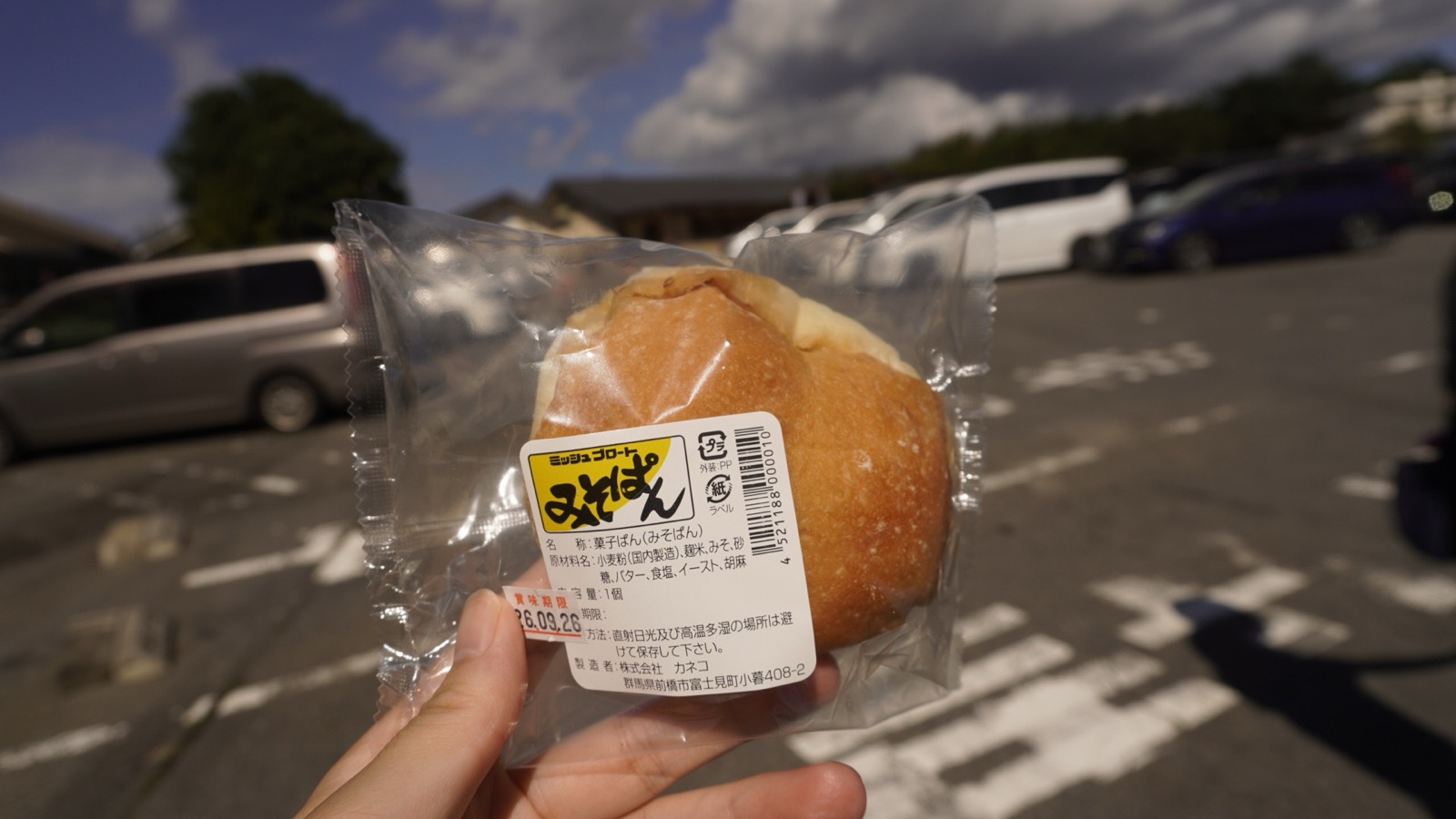 「ミッシュブロート みそぱん」のラベルが貼られた袋入りの丸いパンを、駐車場を背に手で持っている