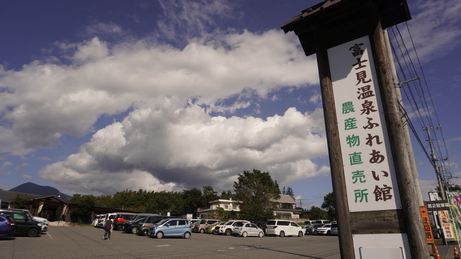 「富士見温泉ふれあい館 農産物直売所」と書かれた木の柱の看板と、車の並ぶ駐車場、その上に広がる雲