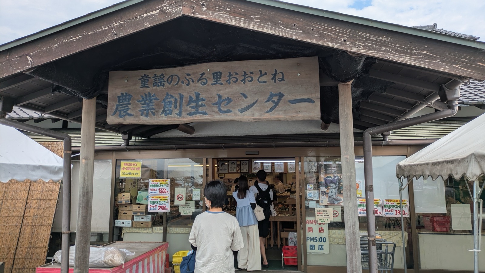 農産物直売所「農業創生センター」の入口。屋根の下に手描きの看板が掲げられ、買い物客が出入りしている