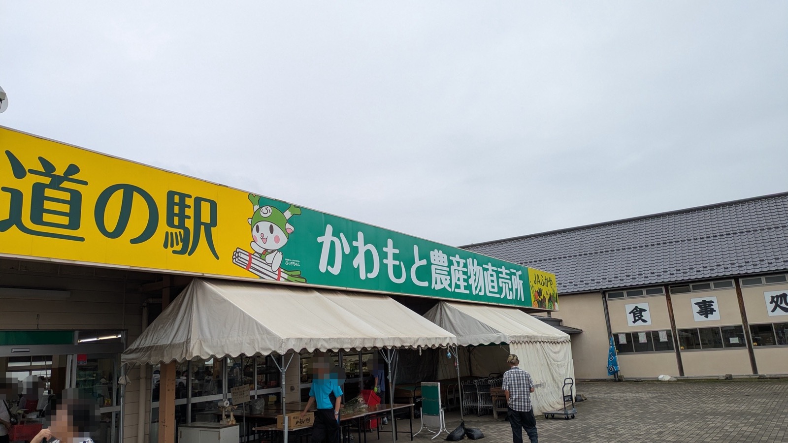 「道の駅」「かわもと農産物直売所」「JAふかや」の看板を掲げた直売所の入口。軒先に白いテントが張られ、右手に「食事処」と書かれた瓦屋根の建物が建つ