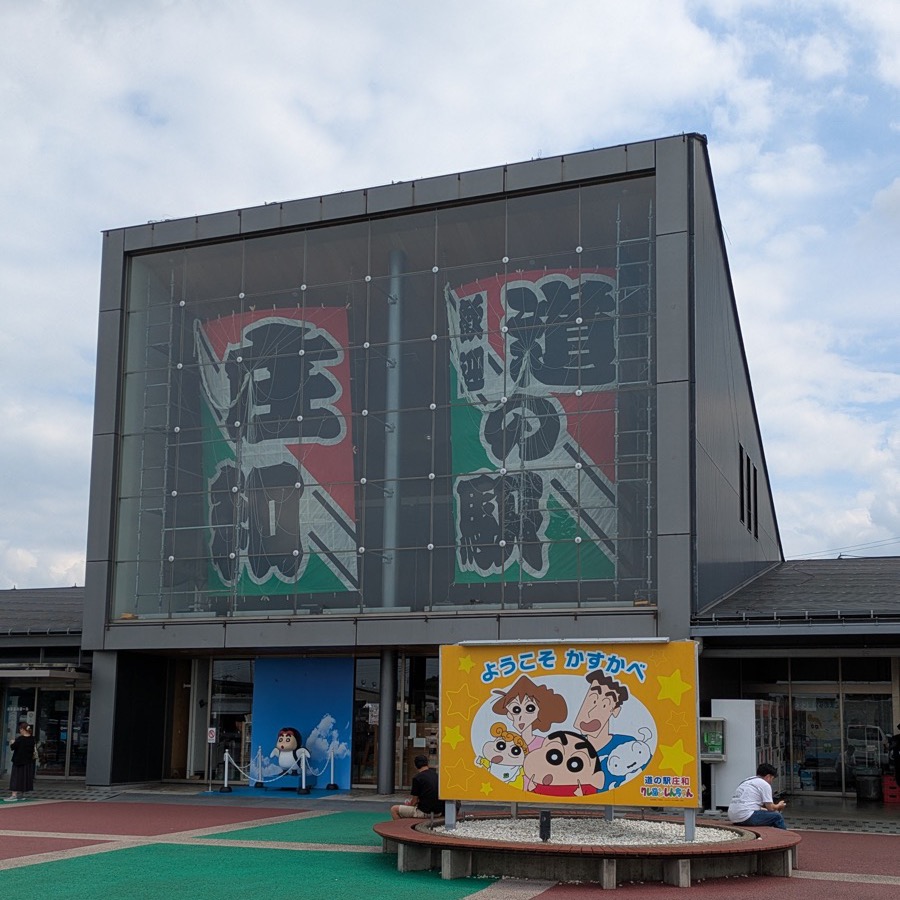 「道の駅 庄和」と書かれた大きな凧が掲げられた建物と、クレヨンしんちゃんの案内看板