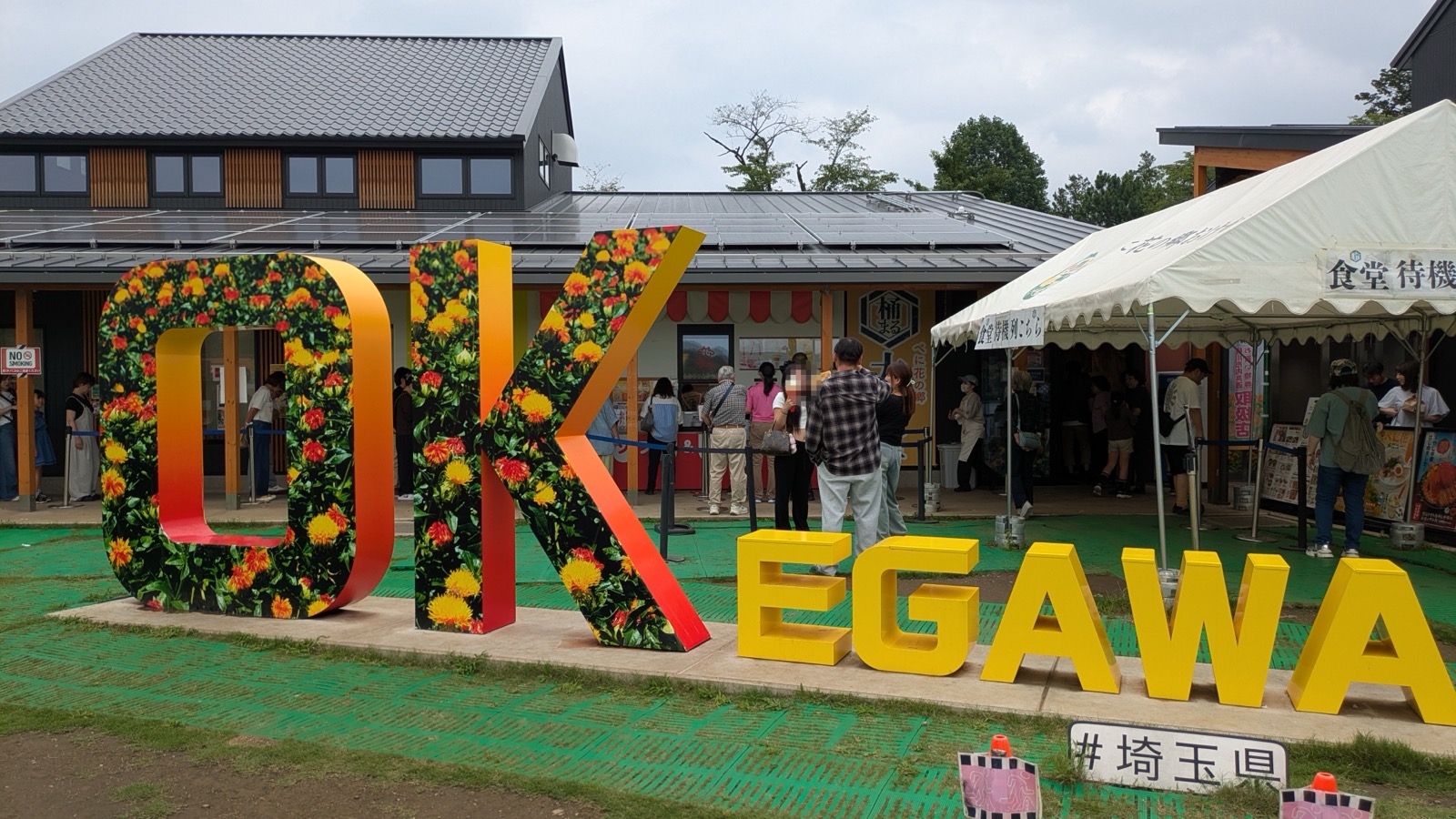 「OK EGAWA」の巨大な立体文字オブジェの前を人々が行き交う道の駅べに花の郷おけがわの入口広場