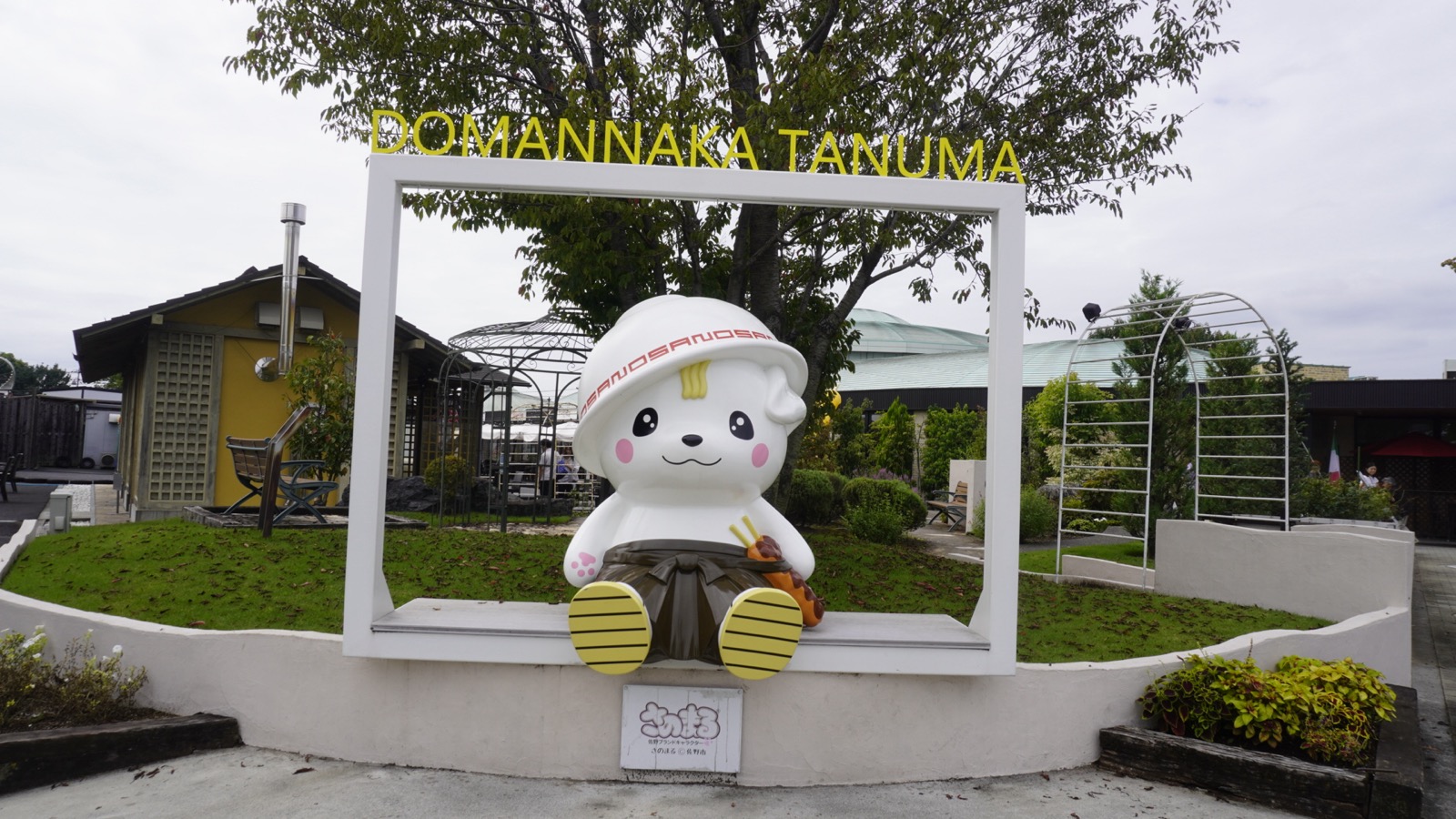 「DOMANNAKA TANUMA」の文字がついた白い写真フレームの中に、佐野らーめんのお椀をかたどった白い笠をかぶり、いもフライの剣を差したさのまるの像が座っている