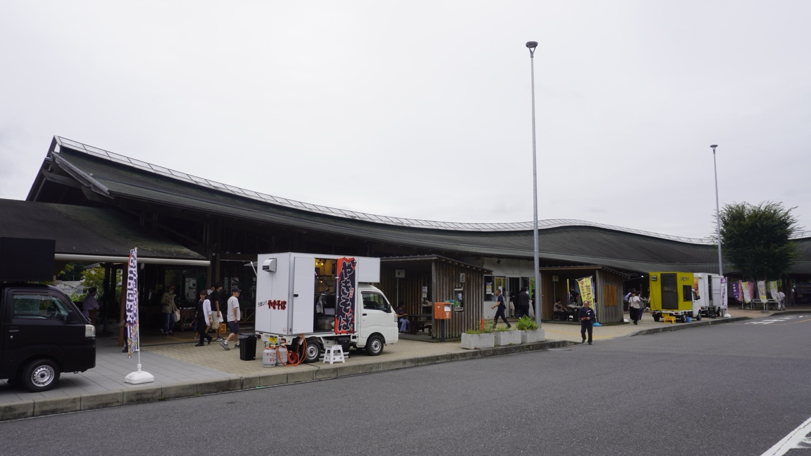 曇り空の下、緩やかに波打つ灰色の大屋根が横に長く続く道の駅みかもの建物。手前の歩道沿いに焼きそばなどのキッチンカーが停まっている