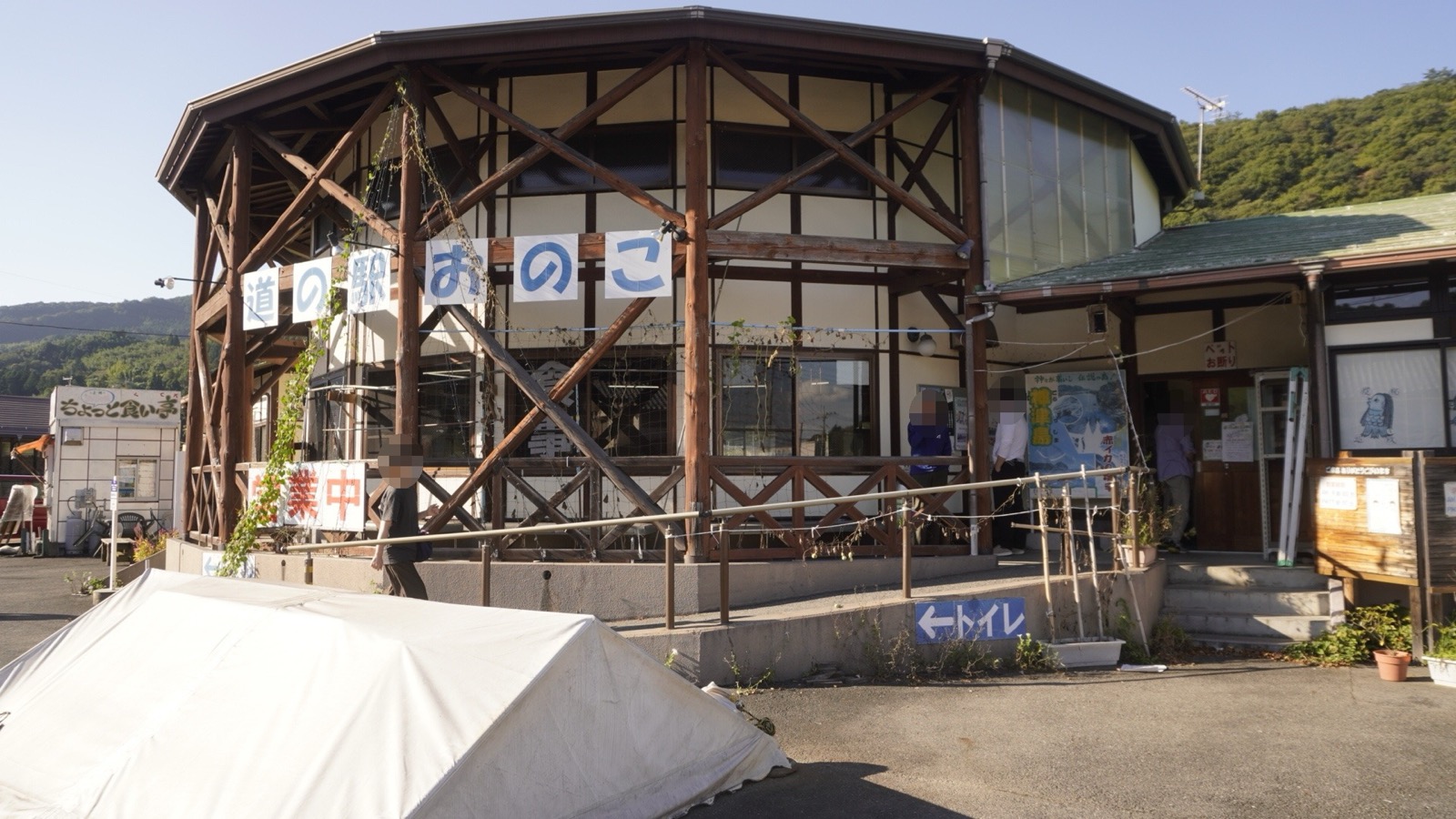 丸太を斜めに組んだ八角形の建物に「道の駅 おのこ」の文字が1字ずつ掲げられ、手前に白いシートを掛けた資材と「←トイレ」の案内板がある