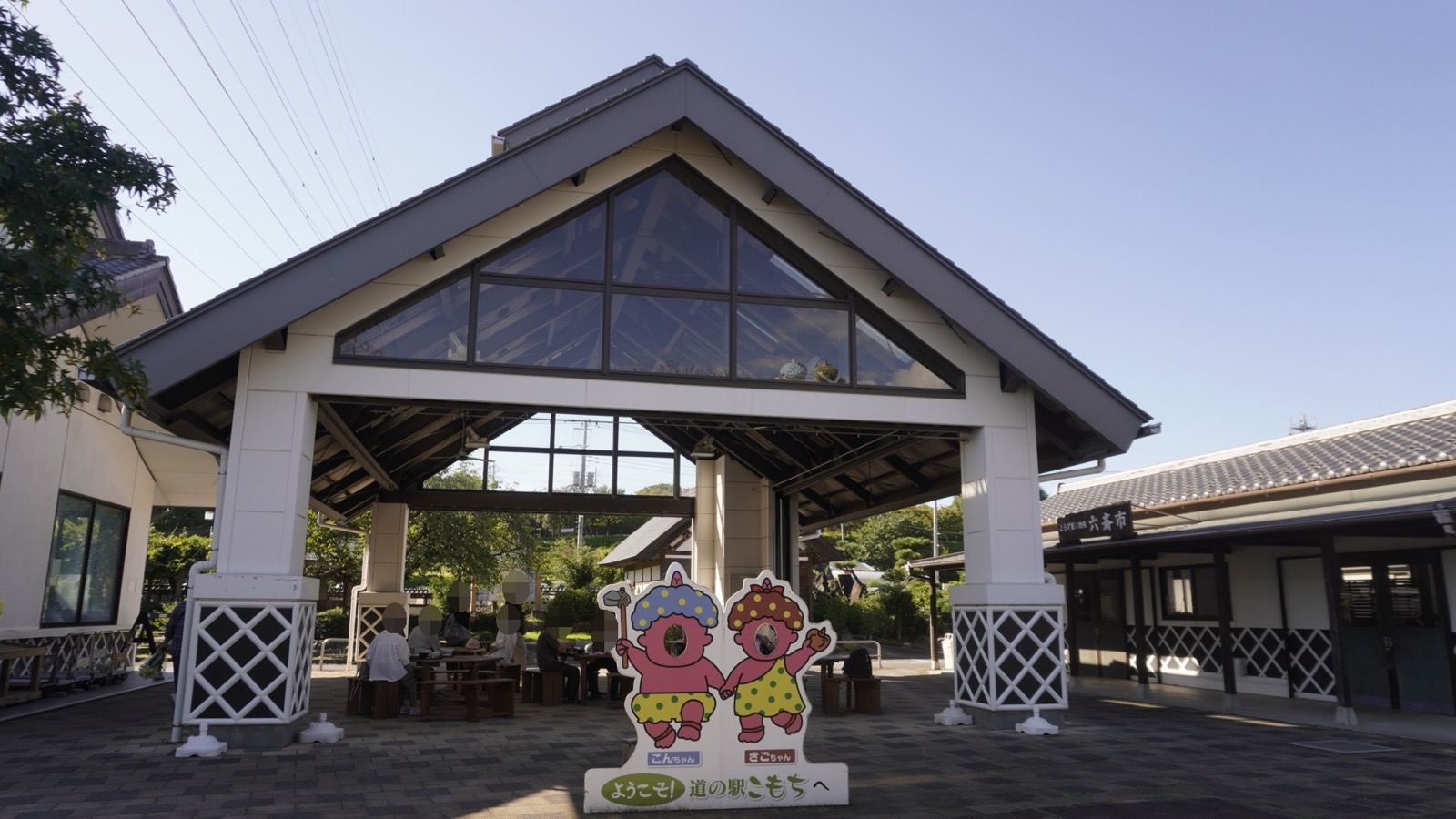 三角屋根の休憩棟の前に「ようこそ!道の駅こもちへ」と書かれた2人組キャラクターの顔出しパネル。奥のテーブルで食事をする人たち
