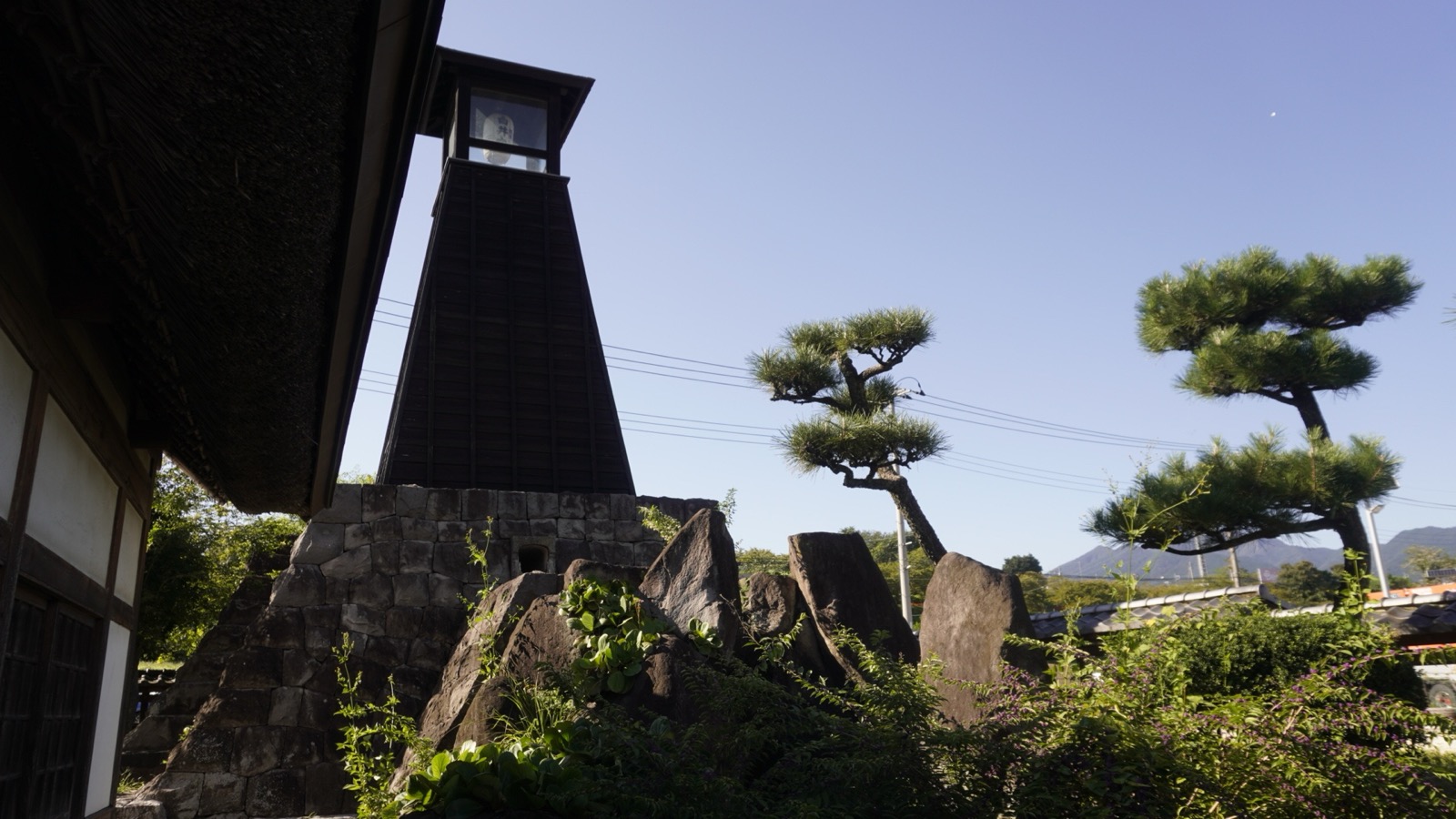 茅葺き屋根の建物の脇、石垣の上に建つ黒い板張りの塔と、手前の岩組みの庭、松の木