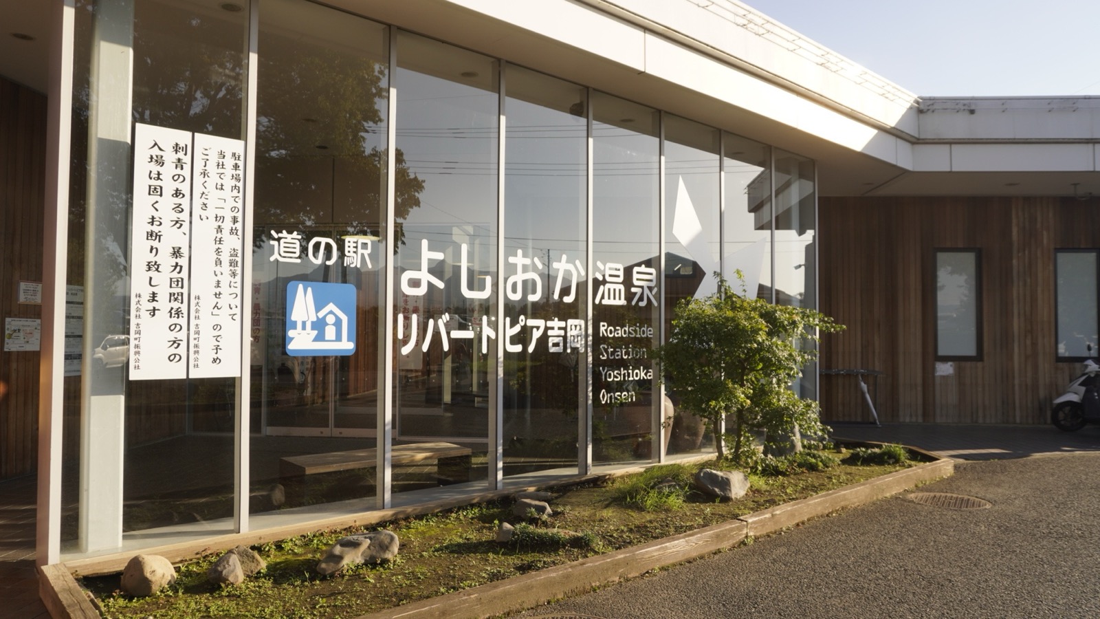 木目の外壁とガラス張りの建物。ガラス面に「道の駅 よしおか温泉 リバートピア吉岡」「Roadside Station Yoshioka Onsen」の文字と道の駅マーク、左の柱に縦書きの貼り紙が2枚ある