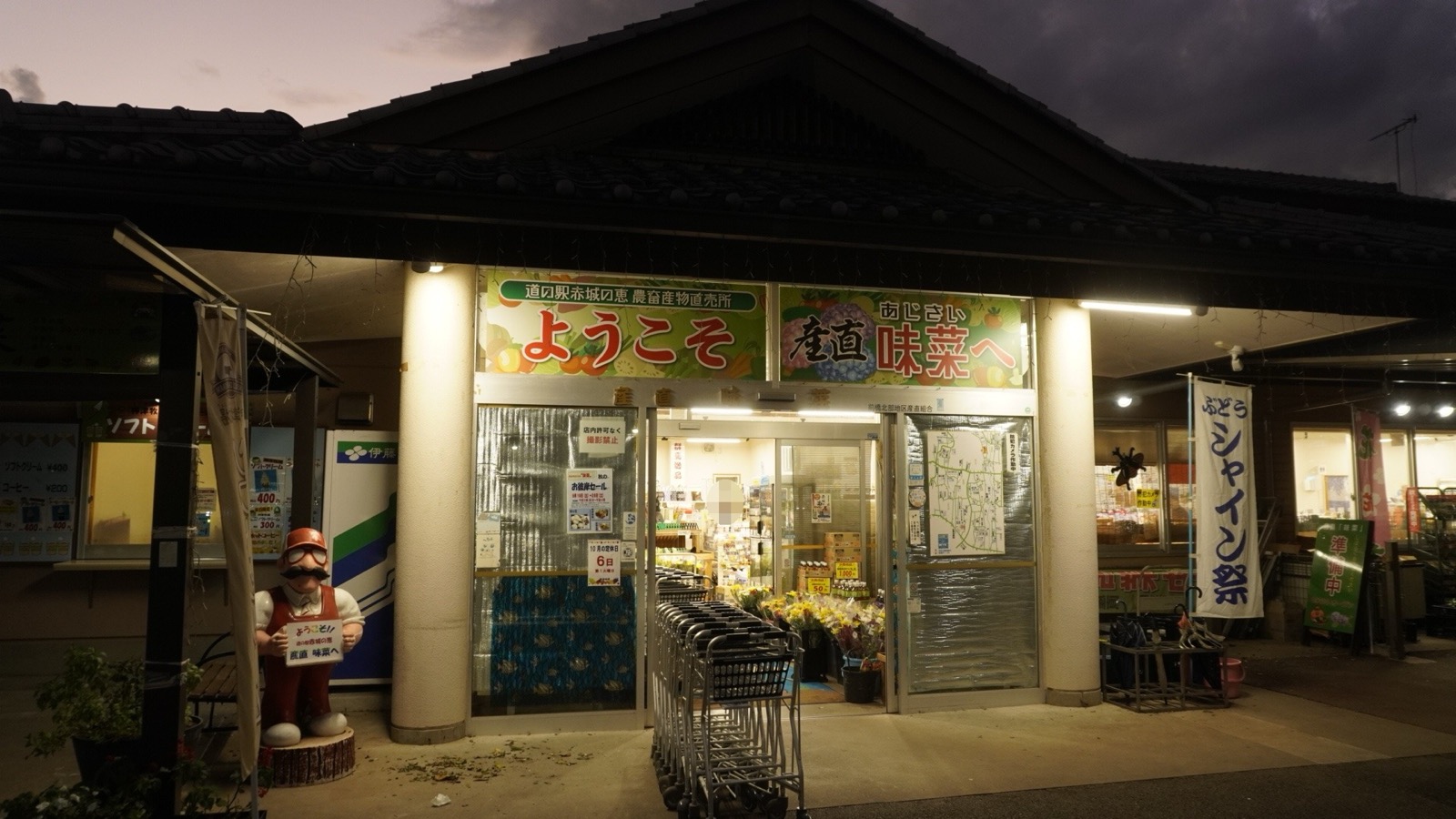 夕暮れの瓦屋根の建物の入口。「道の駅赤城の恵 農畜産物直売所 ようこそ」「あじさい 産直 味菜へ」の看板の下にガラス戸と買い物カートが並び、左に看板を持った人形、右に「ぶどう シャイン祭」ののぼりが立っている