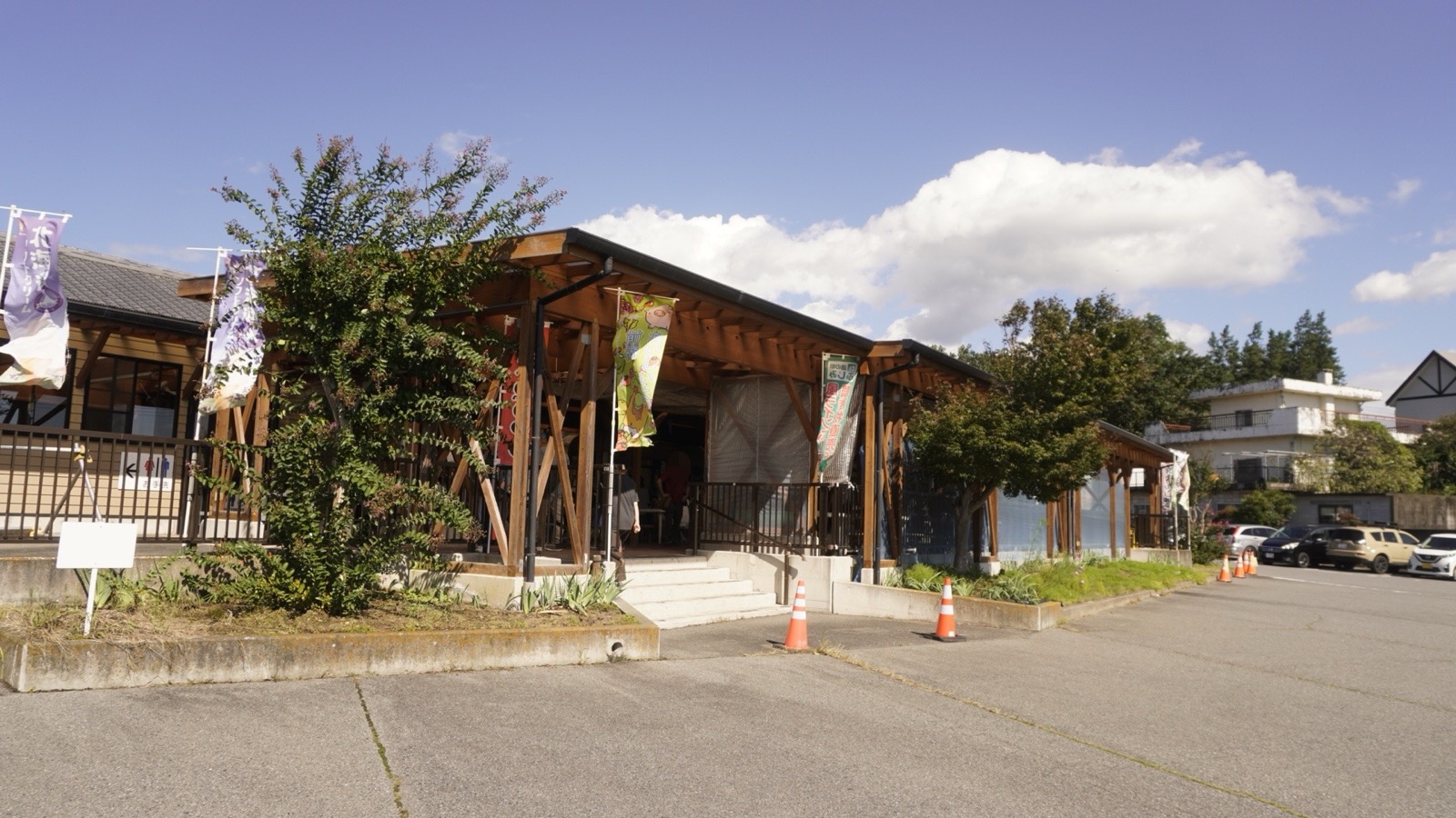 木の柱と梁を組んだ屋根付きの建物の入口に、のぼり旗が並ぶ。手前にサルスベリの木とコーン、右手に駐車場
