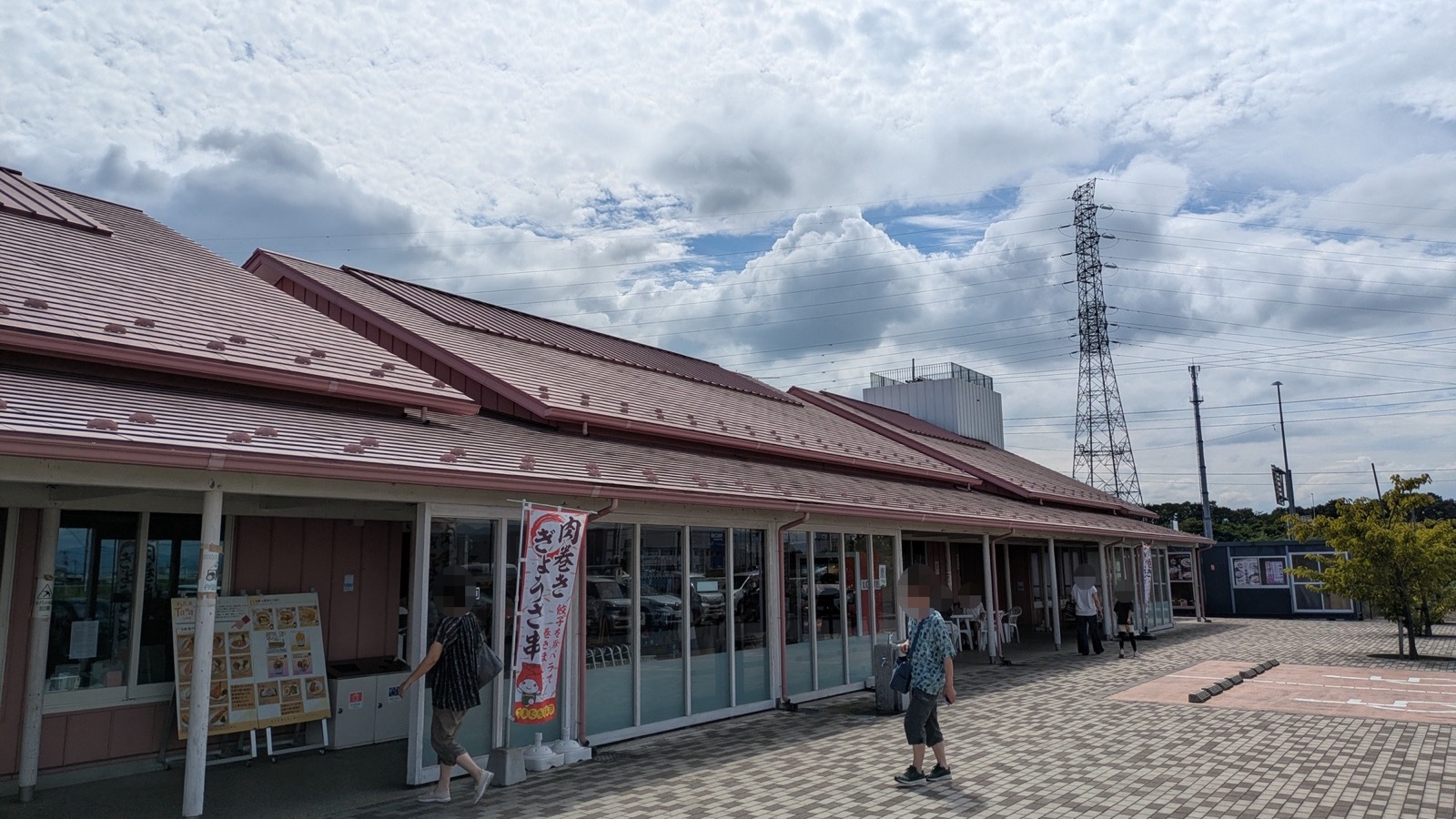 赤茶色の屋根が連なる平屋の建物と、ガラス張りの店先。入口前に「肉巻きぎょうざ串」ののぼり旗とメニュー看板が立ち、建物の奥に送電鉄塔が見える