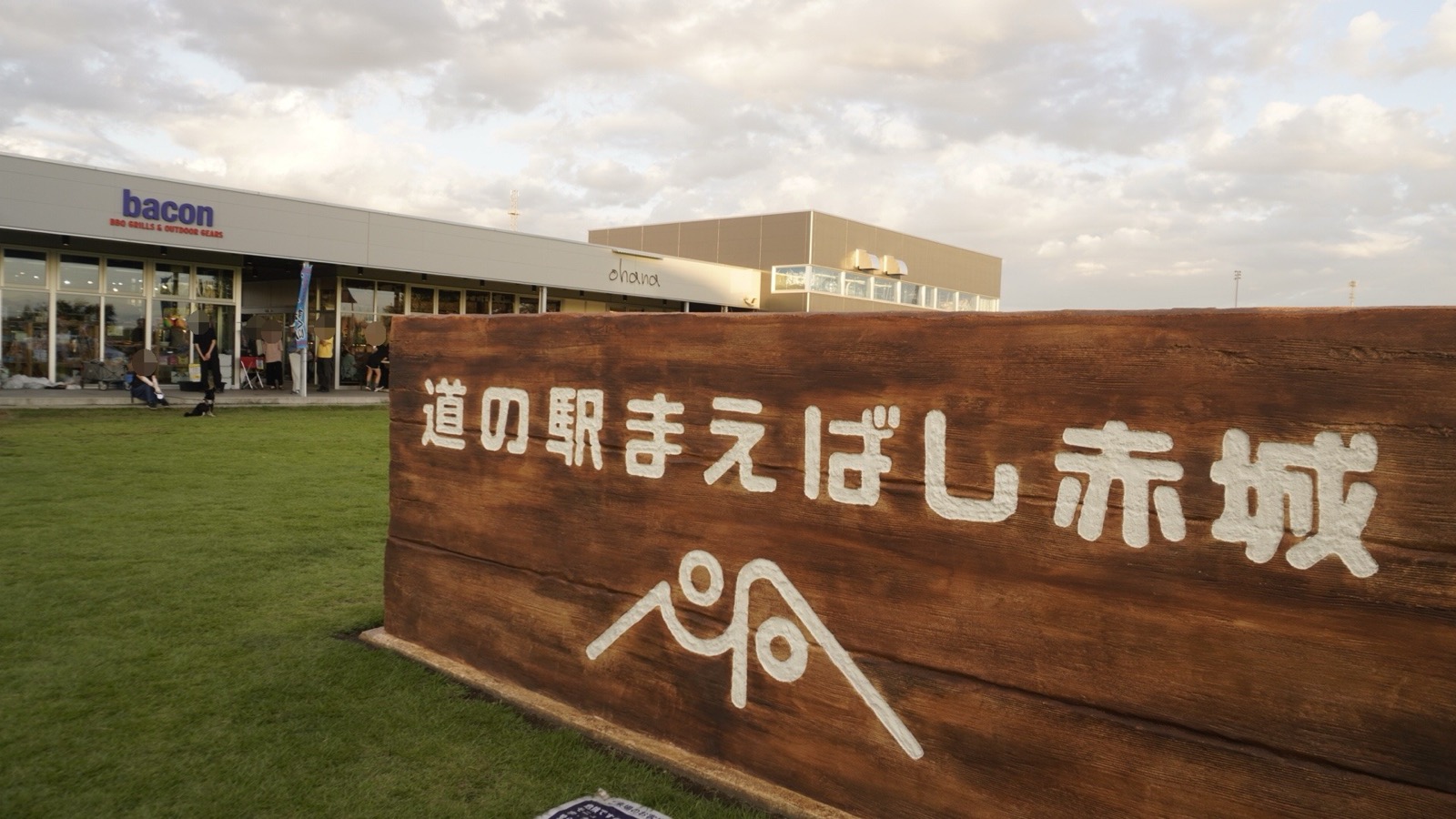 芝生の上に置かれた木目調の大きな看板。白い文字で「道の駅まえばし赤城」と山のロゴが描かれ、左奥に「bacon」「ohana」の看板を掲げたガラス張りの店舗棟がある