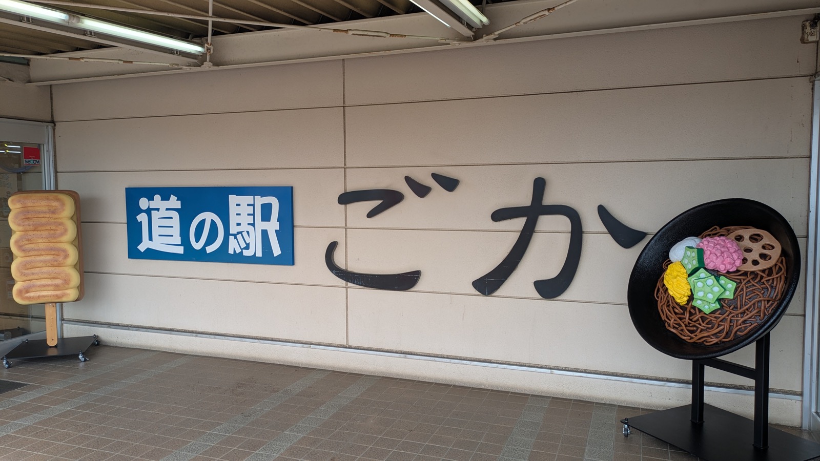 壁に取り付けられた「道の駅 ごか」の看板。左に棒付きの焼き菓子のような形の立て看板、右に野菜をのせた揚げそばの器を模したモニュメントが置かれている