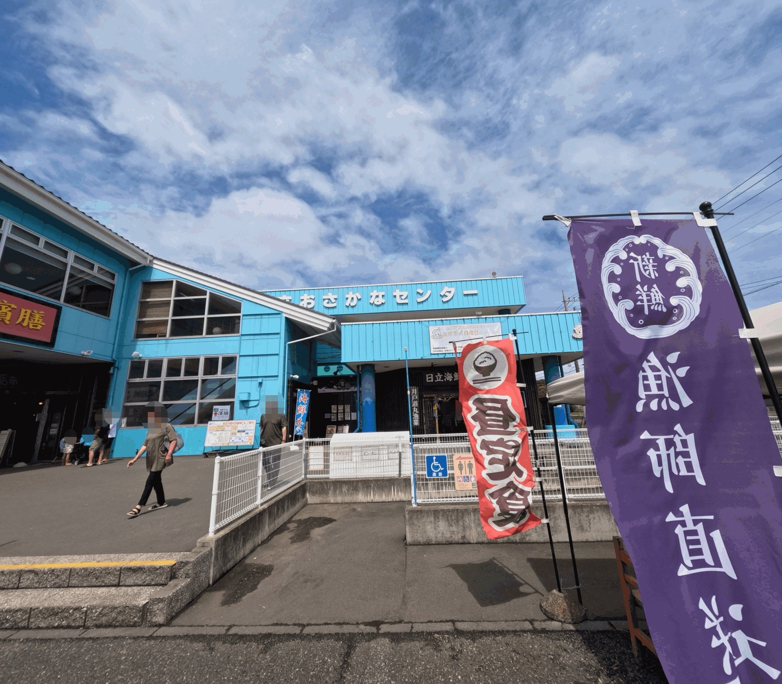 水色の外壁の日立おさかなセンターの建物と入口。手前に「漁師直送」「昼定食」ののぼりが立つ