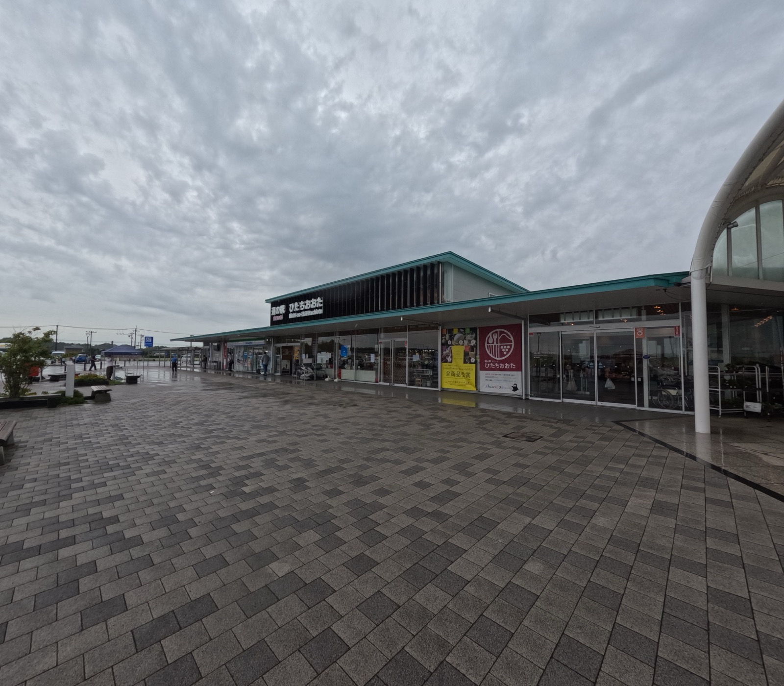 曇り空の下、雨に濡れた石畳の広場の奥に建つ「道の駅 ひたちおおた」の建物。緑の屋根の下にガラス張りの店舗が並び、入口脇に黄色と赤の垂れ幕が下がっている